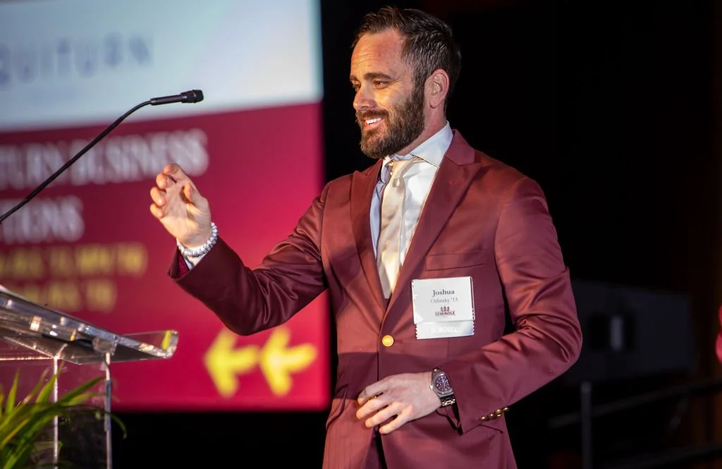 Josh Orlinsky standing at a podium reaching for a microphone and smiling